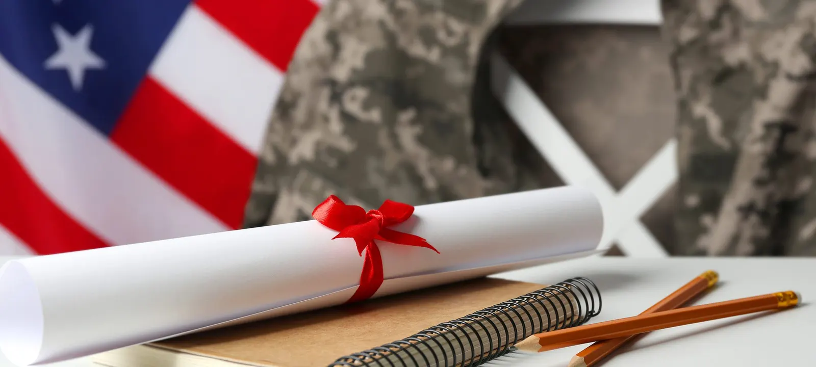 A diploma tied with red ribbon in front of an American flag and a military uniform